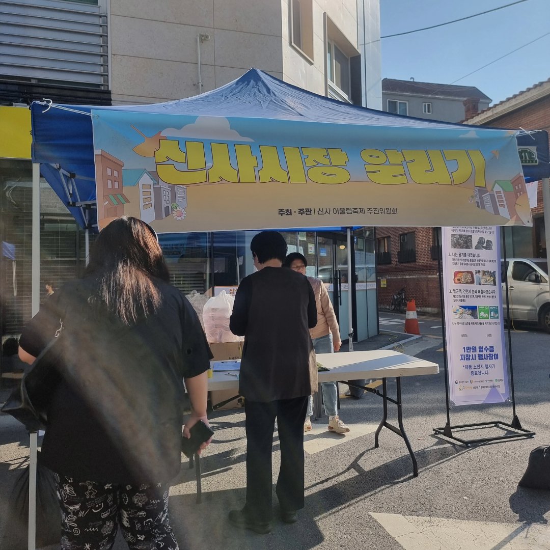 '신사시장 알리기' 현수막을 단 파란 천막 부스 앞에 사람들이 등을 보이고 서 있다