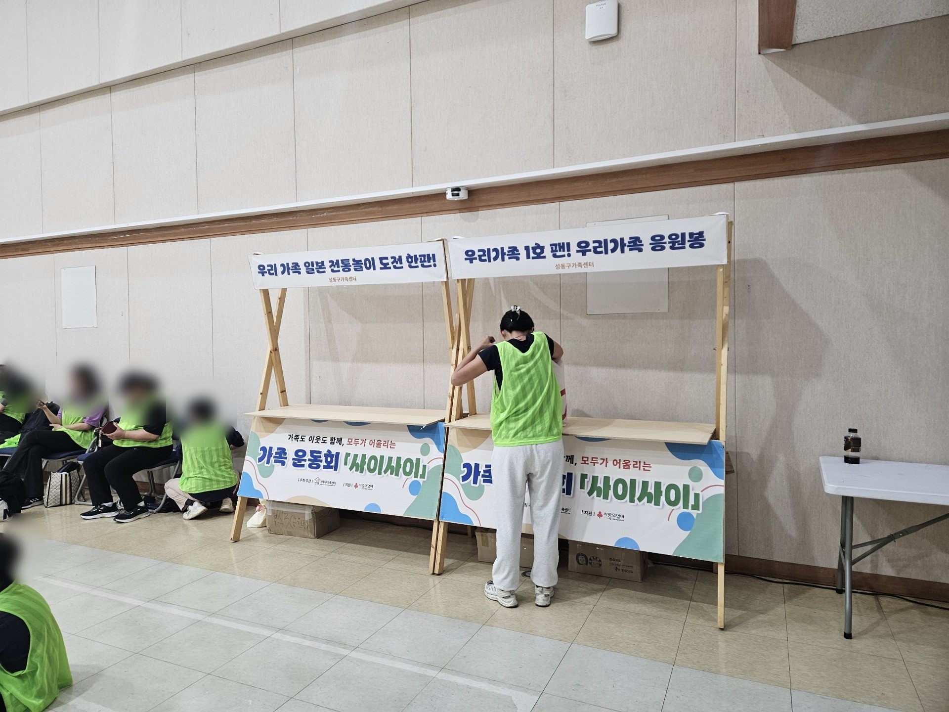 대강당 벽을 따라 일본 전통놀이와 응원봉 만들기 부스의 나무 X매대가 나란히 서 있다. 인물 얼굴은 흐리게 처리했다