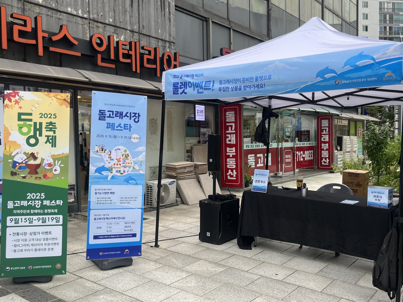 2025 동행축제 배너와 돌고래시장 페스타 배너가 롤렛이벤트 캐노피 옆에 서 있고 테이블과 스피커가 놓여 있다
