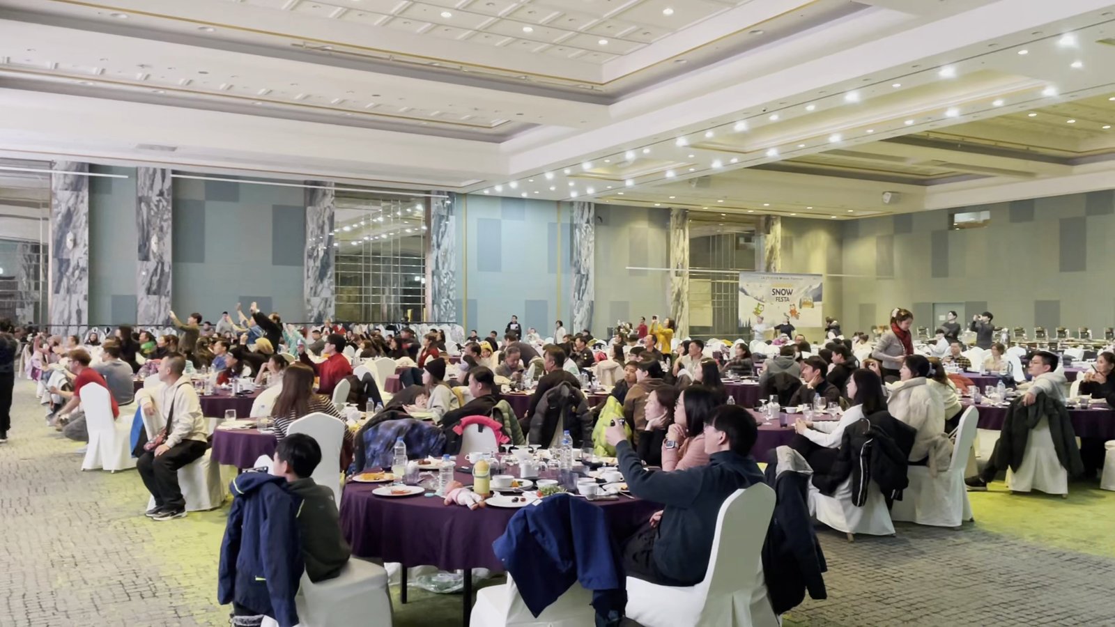 흰 의자와 라운드 테이블이 가득한 연회장에서 관광객들이 식사하며 앞쪽 무대를 보고 있다