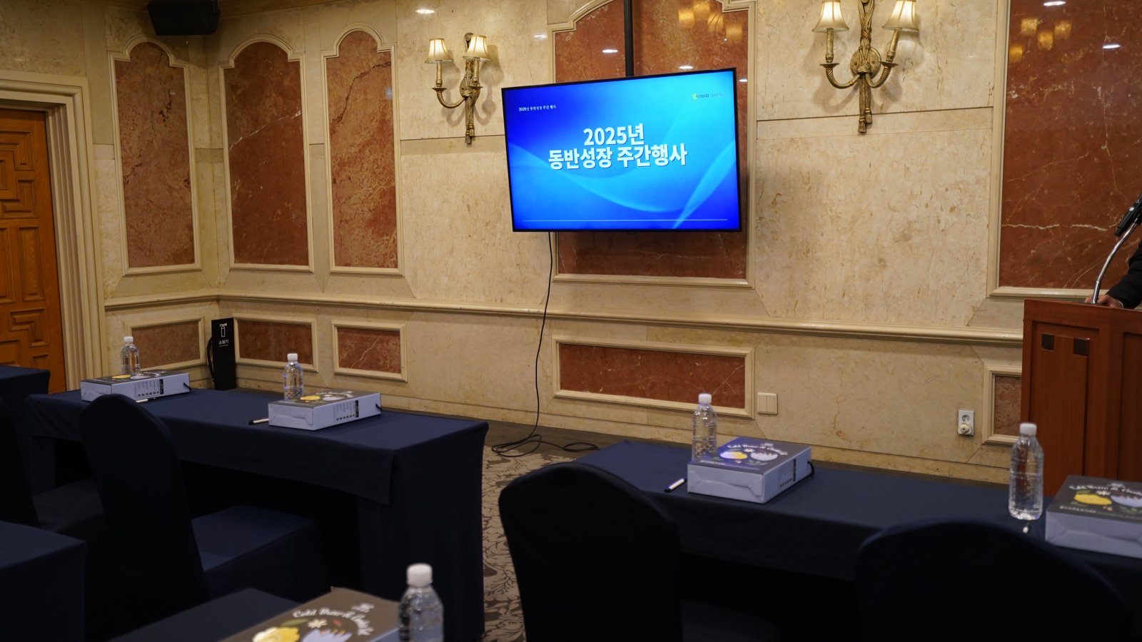 대리석 벽에 걸린 모니터에 2025년 동반성장 주간행사 화면이 떠 있고 검은 천을 씌운 테이블마다 물병과 자료 상자가 놓여 있다