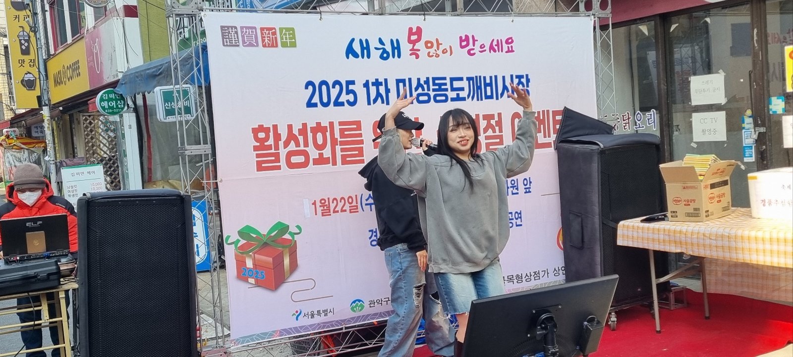 현수막 무대 앞에서 댄서 두 명이 팔을 들어 객석을 향해 인사하고 있고 오른쪽에 경품 테이블과 상자가 보인다