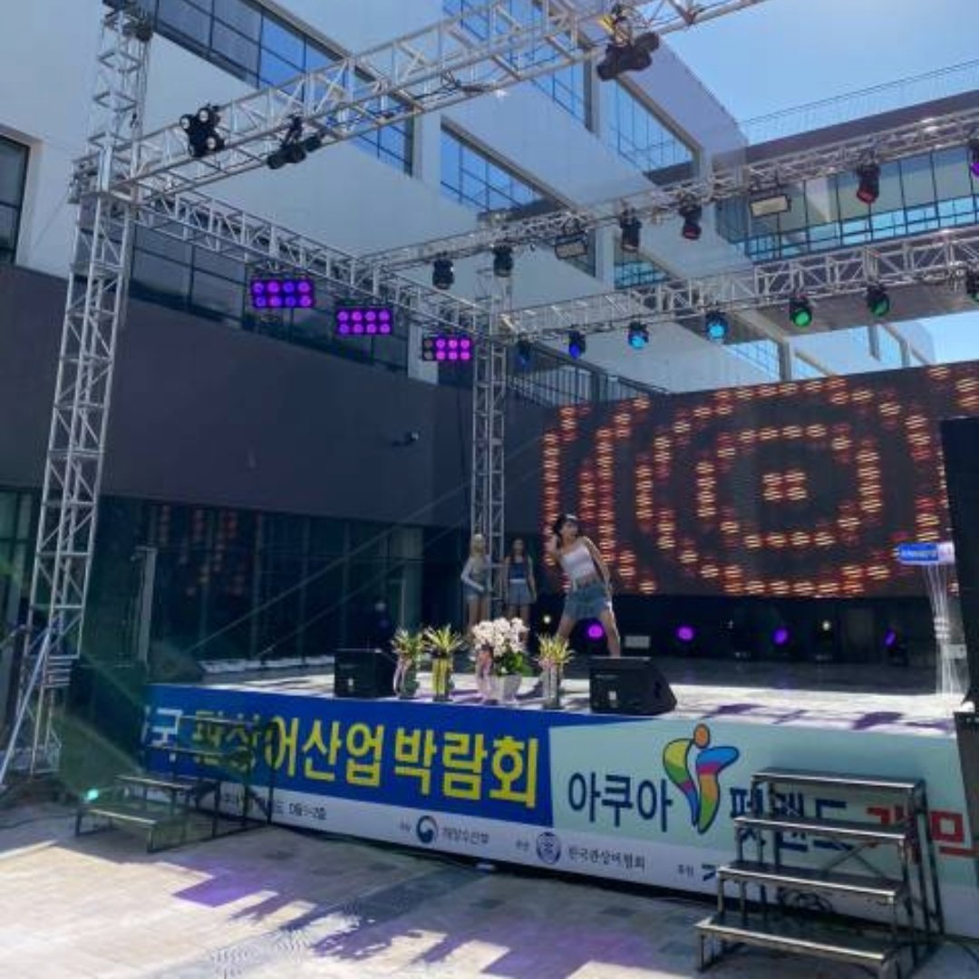 대낮의 야외 무대 위 LED 화면에 붉은 패턴이 떠 있고 그 앞에 출연자가 서 있다