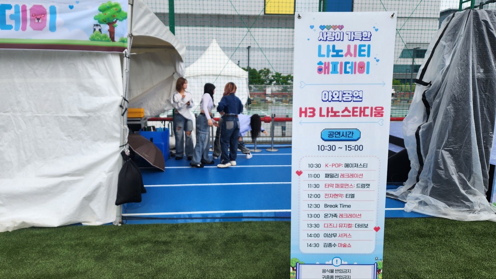 인조잔디 경기장 가장자리에 투명 비닐을 씌운 스피커와 카메라가 서 있고 우비를 입은 촬영자가 카메라를 조작하며 뒤로 흰 천막들이 이어진다