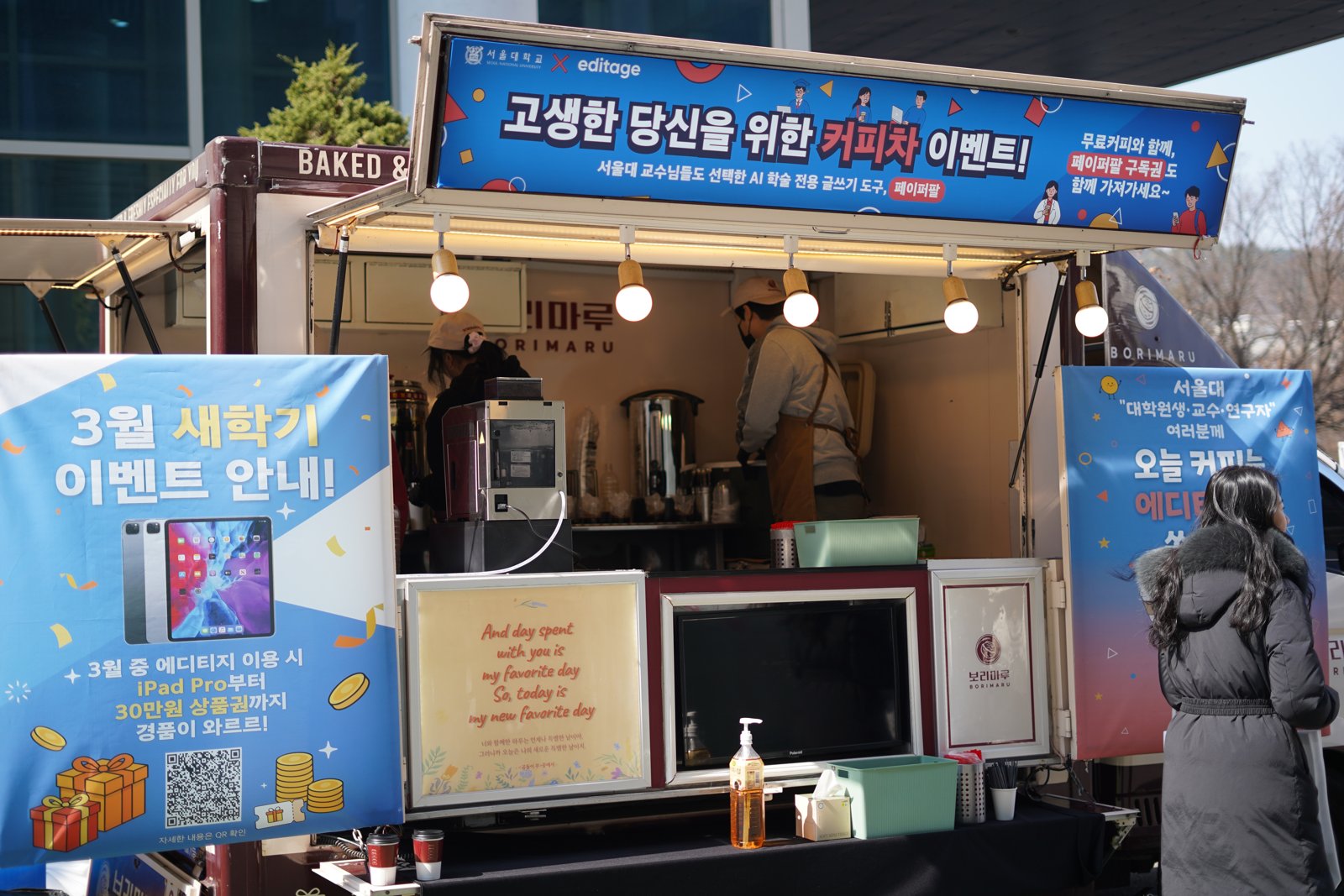 커피차 창구 왼쪽에 아이패드 프로 그림과 QR코드를 넣은 3월 새학기 이벤트 안내판이 서 있고 창구 안에서 바리스타가 커피를 준비하고 있다