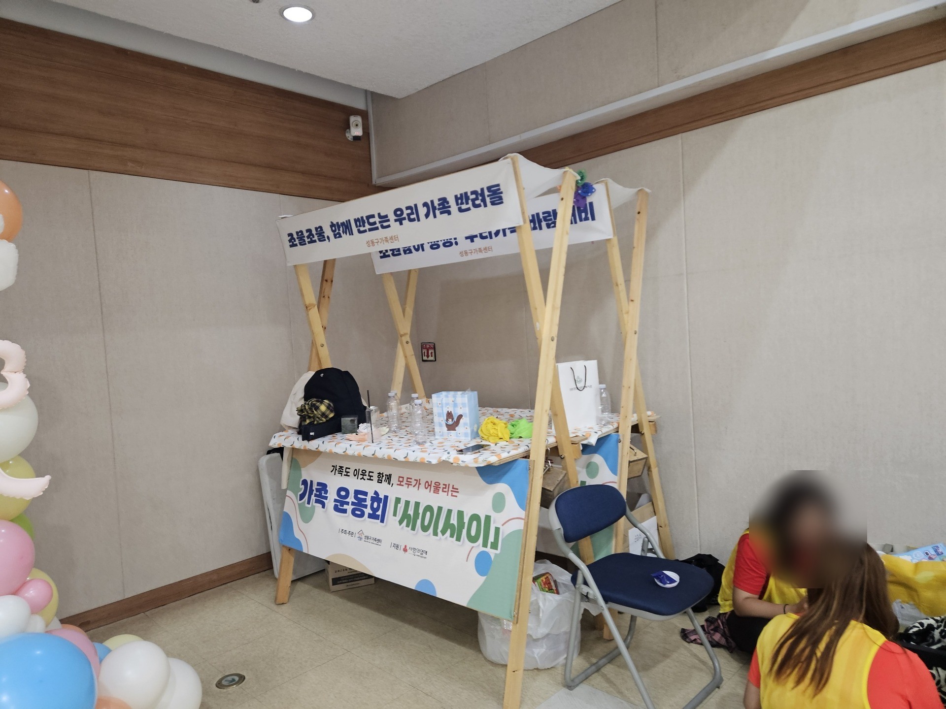 각목을 X자로 엇갈려 세운 나무 매대 위쪽에 반려돌 만들기 부스 현수막, 아래쪽에 가족 운동회 사이사이 현수막이 붙어 있다. 인물 얼굴은 흐리게 처리했다