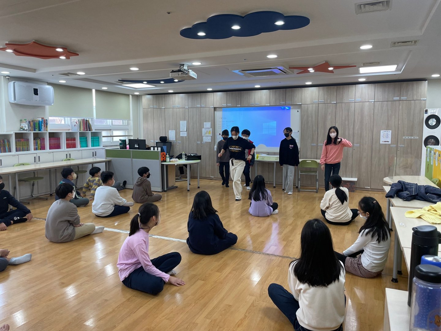 교실 바닥에 앉은 학생들이 앞쪽 화면에 나오는 영상을 보고 있다