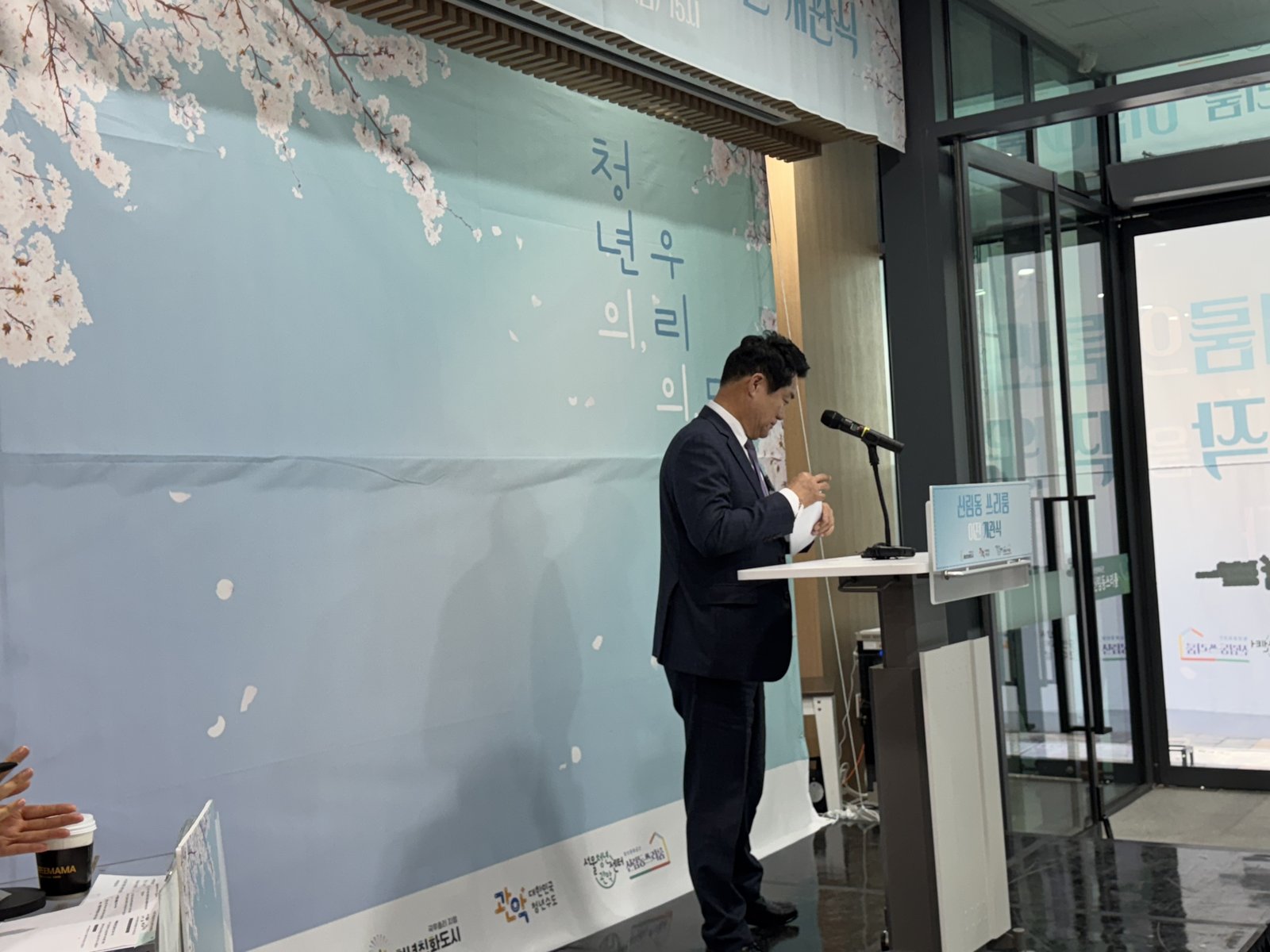 벚꽃 그림 배경막 앞 단상에서 정장 차림의 내빈이 축사를 하고 있다