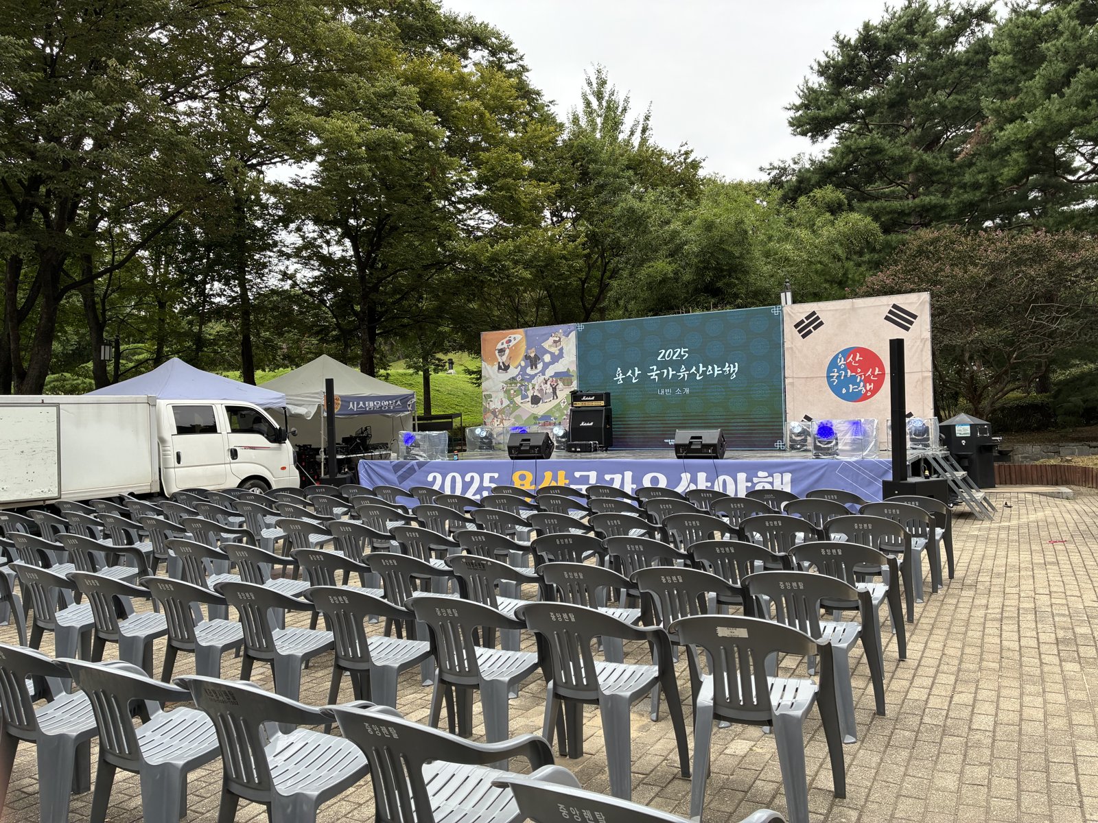 무대 앞 광장에 회색 의자가 넓게 깔리고 왼쪽에 트럭과 시스템 운영 천막, 오른쪽에 흰 천막이 서 있다