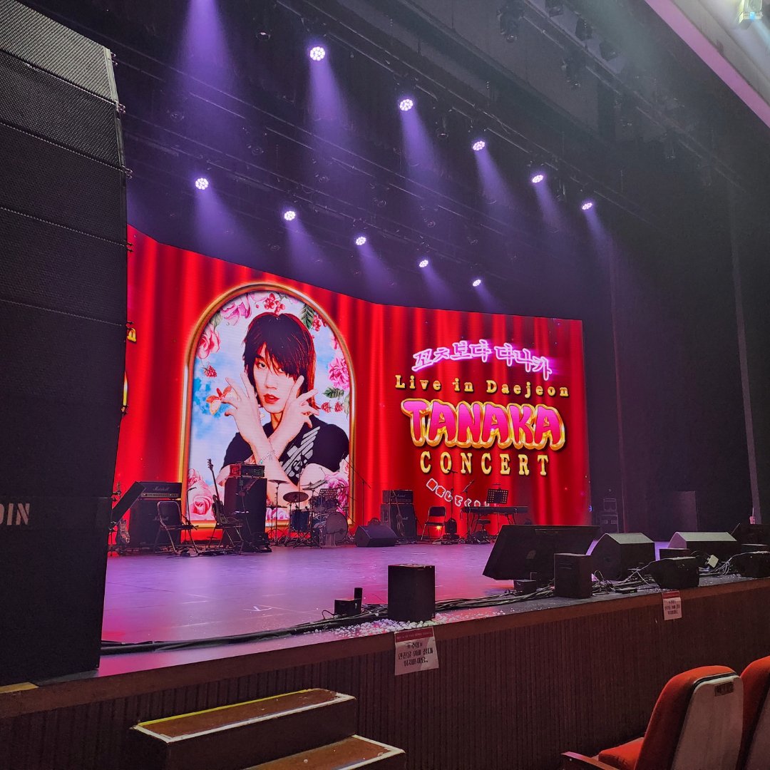 보라색 조명이 켜진 무대 LED에 다나카 콘서트 대전 공연 로고가 떠 있고 앞에는 비어 있는 붉은 객석이 보인다