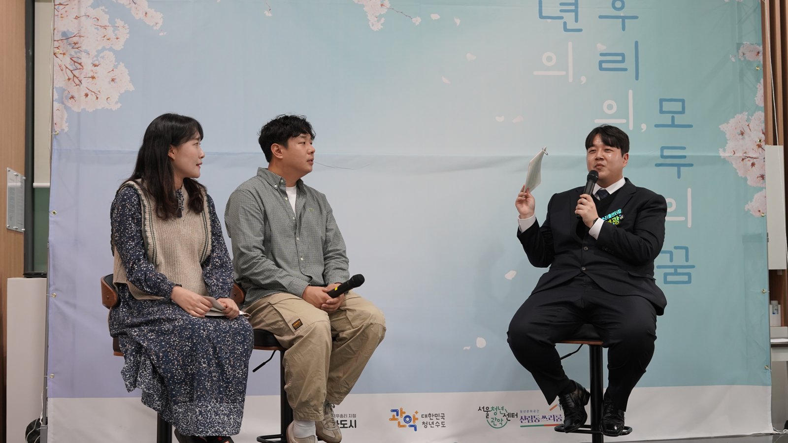 벚꽃 그림 배경막 앞 무대에 세 사람이 높은 의자에 앉아 있고 오른쪽 사람이 마이크와 큐카드를 들고 이야기하고 있다
