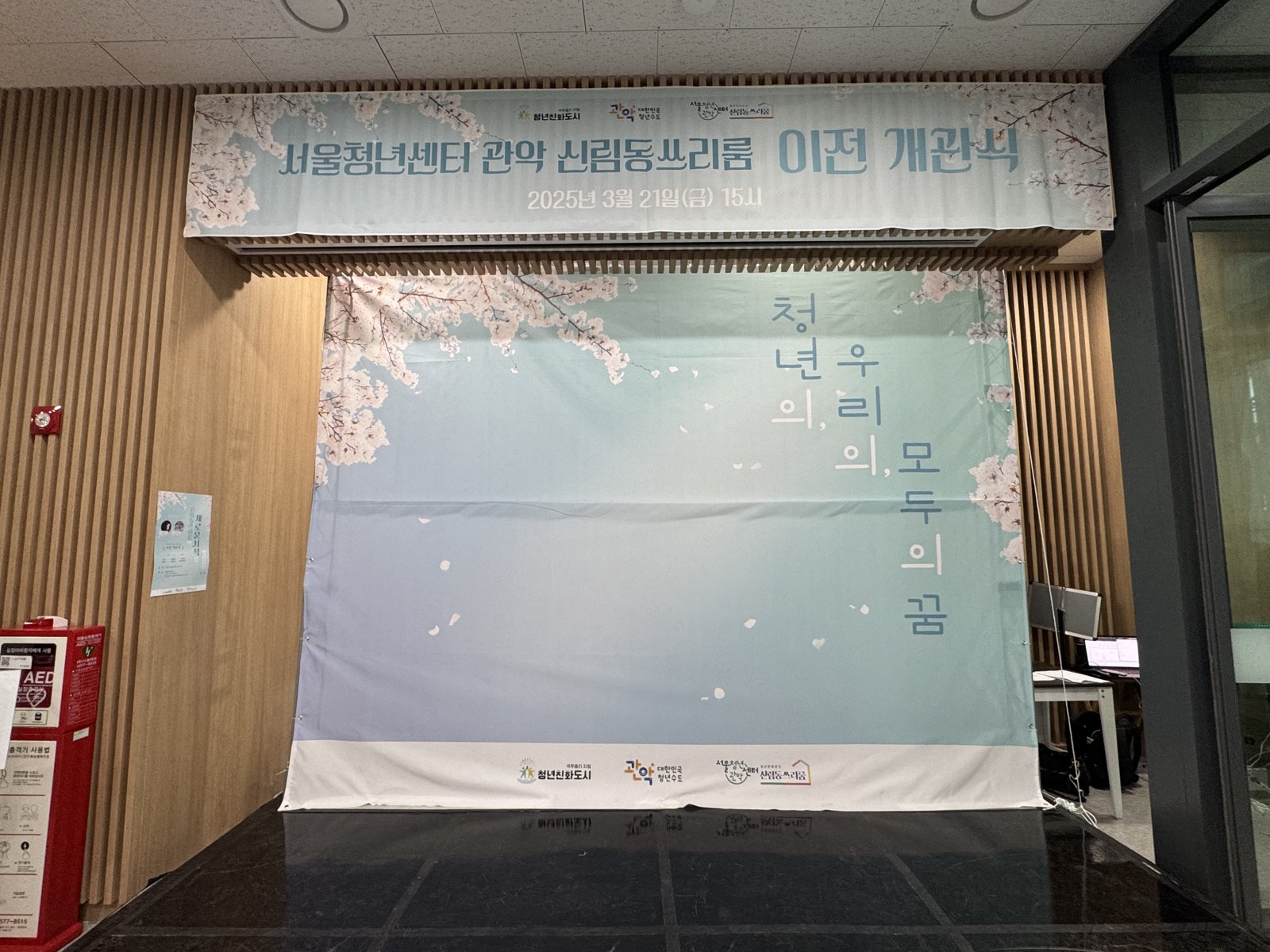 벚꽃 그림 위에 서울청년센터 관악 신림동쓰리룸 이전 개관식 문구와 세로 글씨가 적힌 배경막을 세운 빈 무대