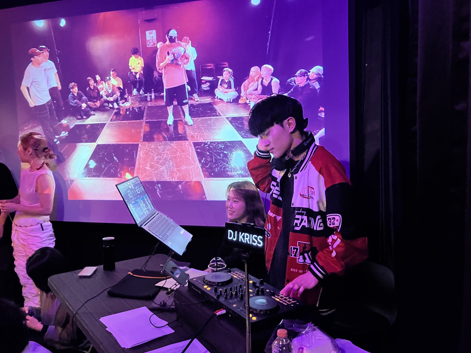 보라색 조명 아래 DJ가 헤드폰을 끼고 믹서를 조작하고 뒤 스크린에 배틀을 지켜보는 참가자들이 비친다
