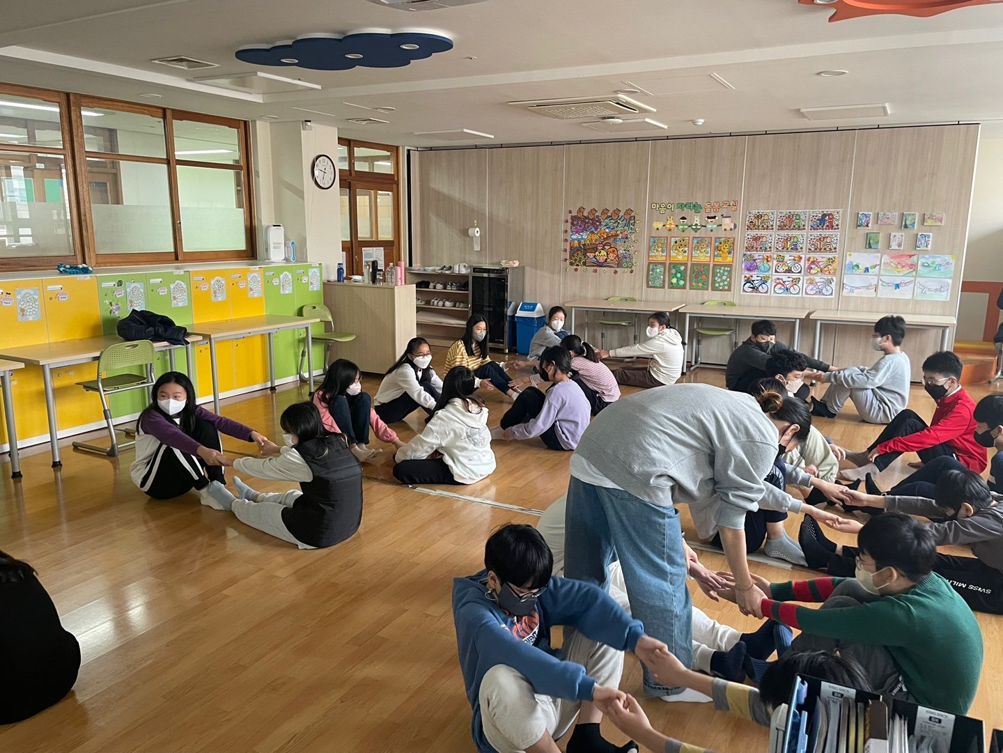 교실 바닥에 앉은 학생들이 둘씩 짝을 지어 다리를 뻗고 스트레칭을 하고 있다