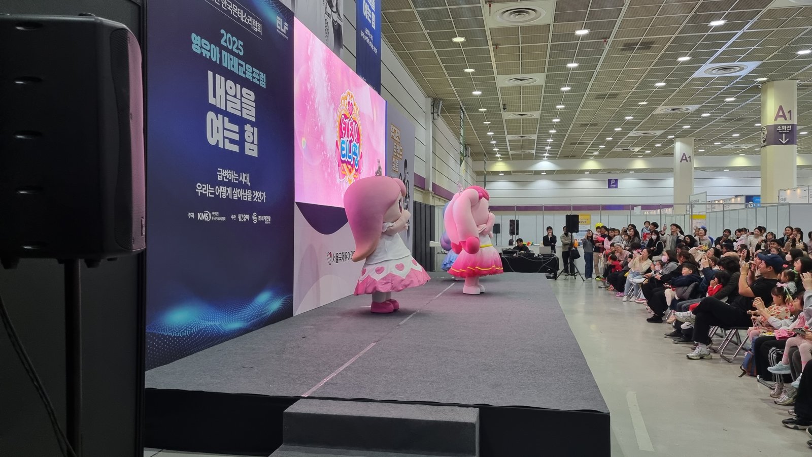 전시장 안 무대에서 분홍색 티니핑 인형탈이 공연하고 무대 옆으로 가족 관객들이 줄지어 앉아 있다