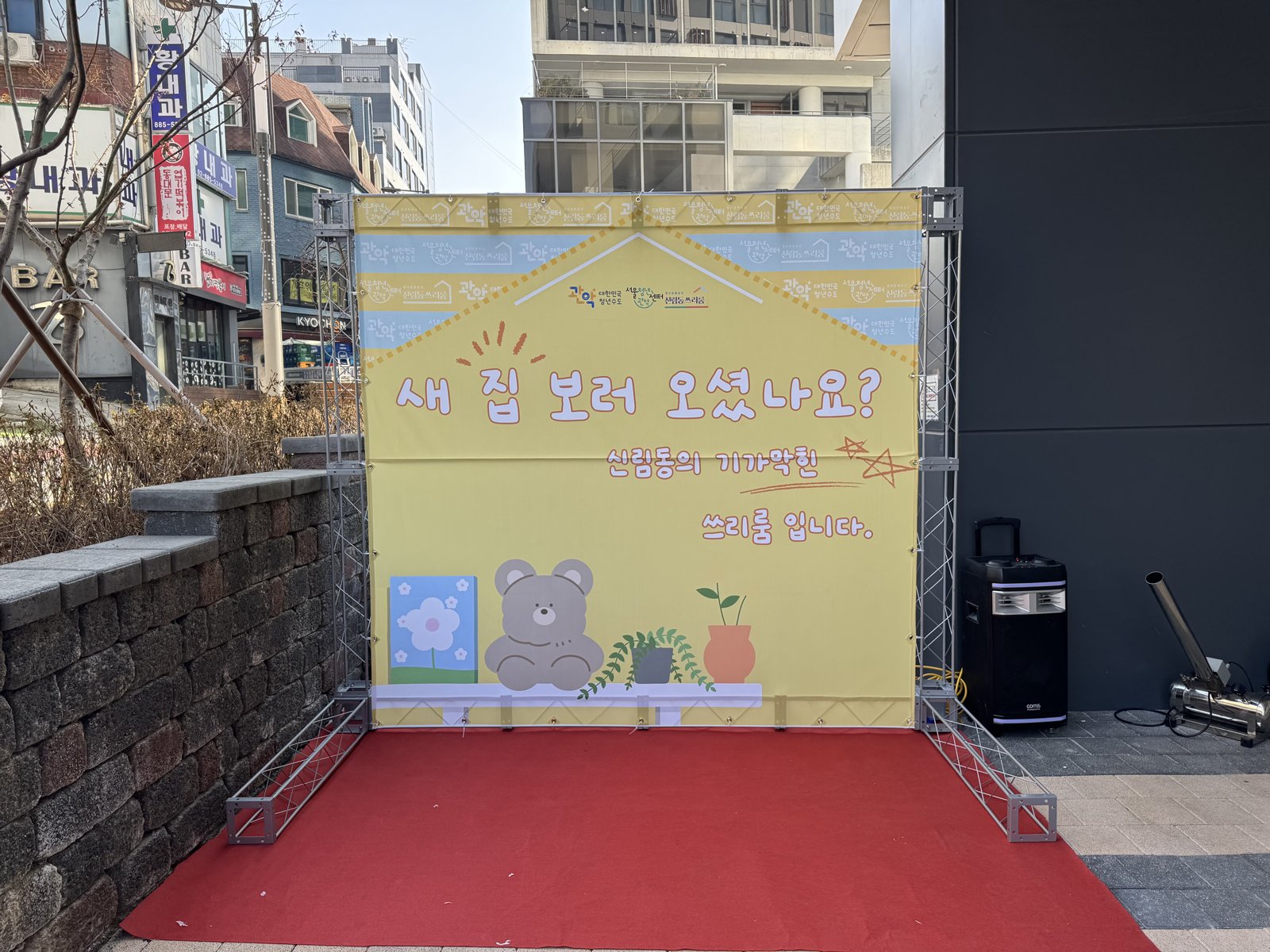 건물 앞 인도에 트러스로 세운 노란 포토월에 '새 집 보러 오셨나요? 신림동의 기가 막힌 쓰리룸입니다' 문구가 적혀 있고 아래에 레드카펫, 옆에 스피커와 에어샷 장비가 놓여 있다