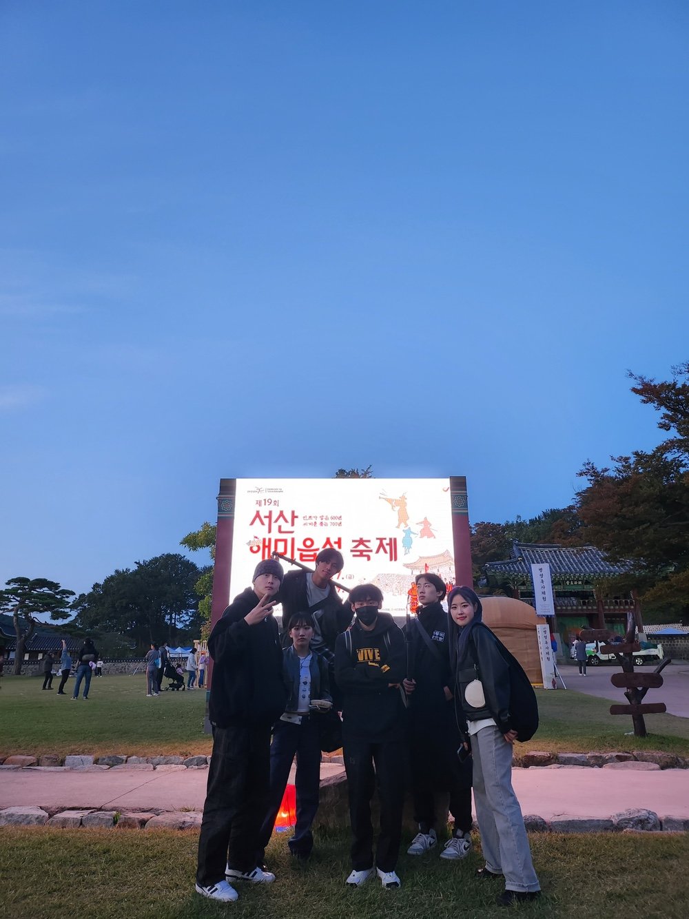해질녘 잔디밭의 축제 안내판 앞에 팀원들이 모여 서 있다