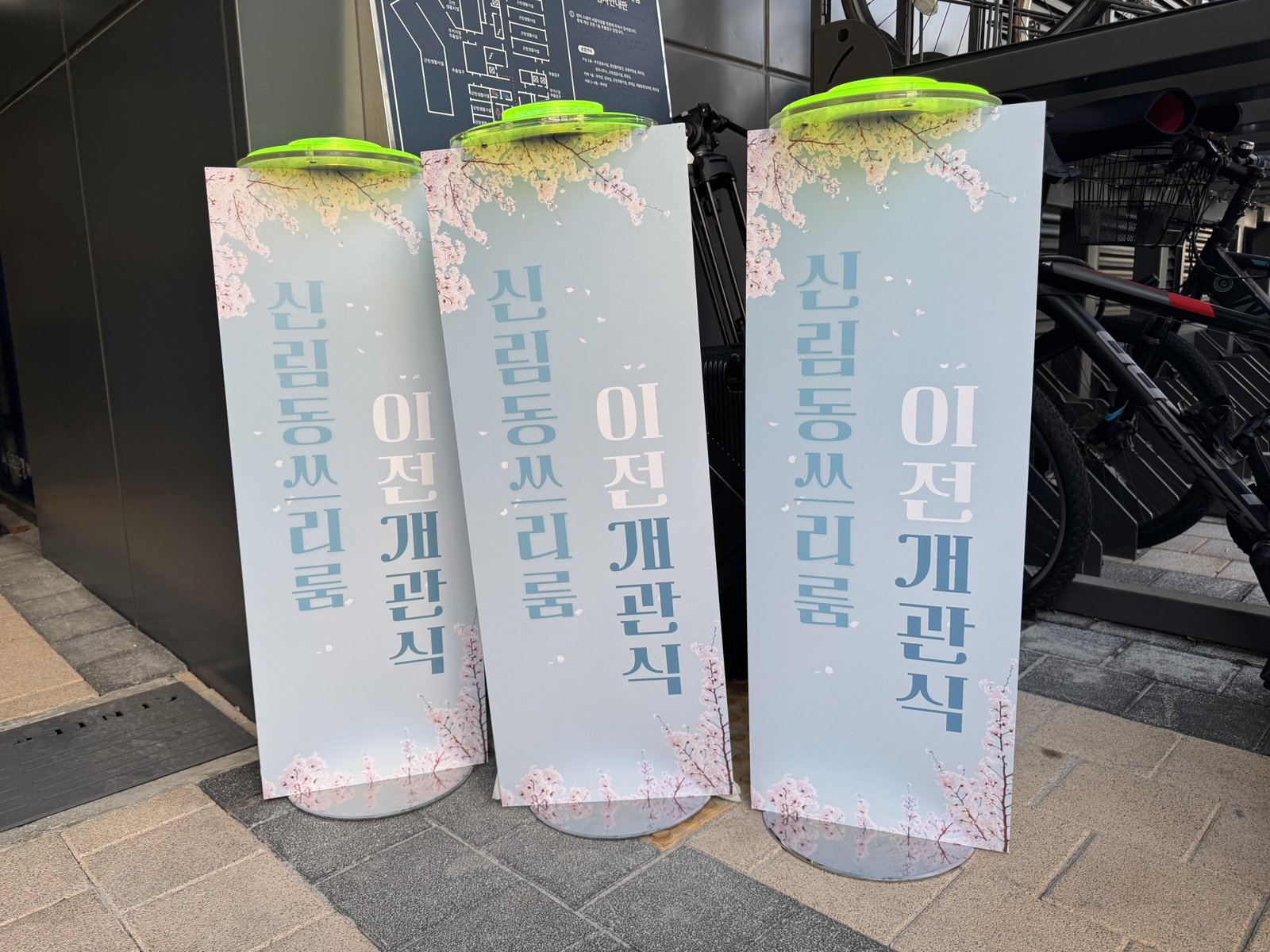 벚꽃 그림과 '신림동쓰리룸 이전 개관식' 문구가 적힌 세로 기둥 배너 세 개가 나란히 서 있고 윗부분에 초록색 버튼이 달려 있다