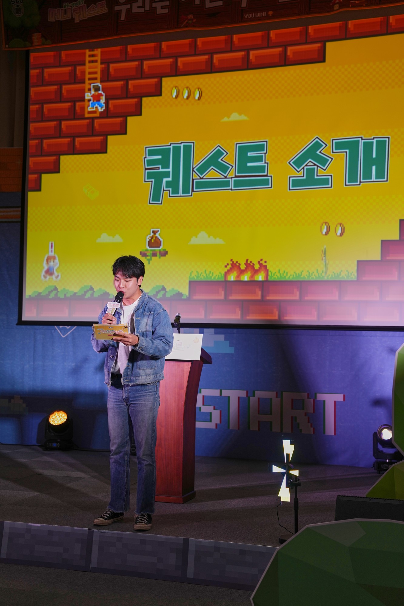 픽셀 게임 화면처럼 꾸민 퀘스트 소개 스크린 앞 연단에서 진행자가 마이크를 들고 말하고 있다
