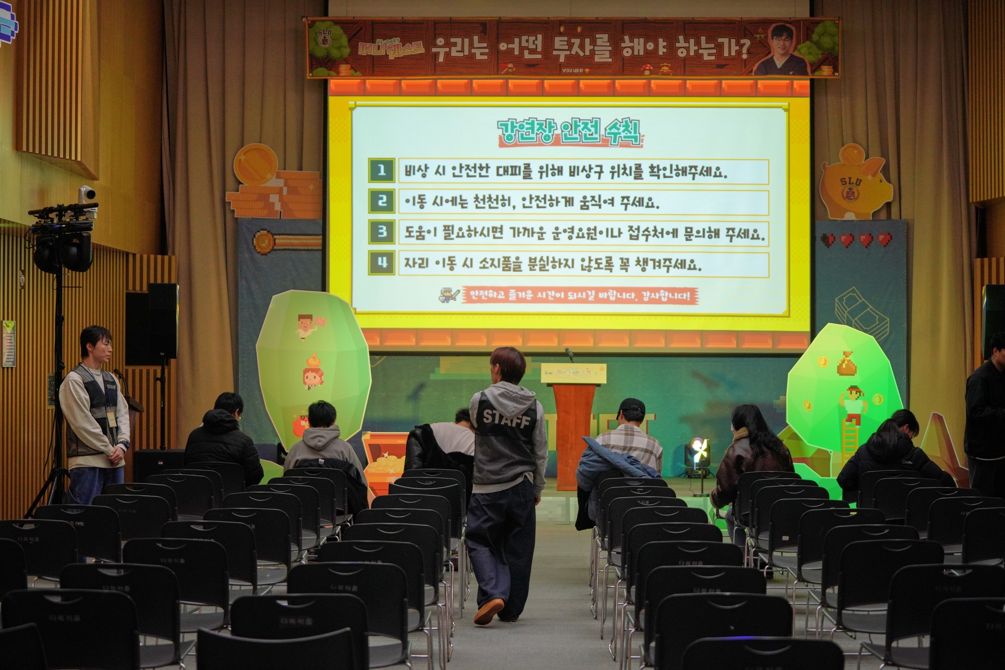 머니 퀘스트 현수막과 스크린, 양옆의 나무·동전 모양 픽셀 조형물이 놓인 회의실 앞쪽과 객석에 앉은 참가자들의 뒷모습