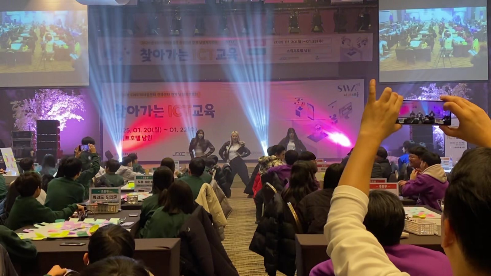 보라색과 파란색 조명이 비추는 볼룸 무대에서 댄서들이 공연하고 객석의 학생들이 뒷모습으로 무대를 보고 있다