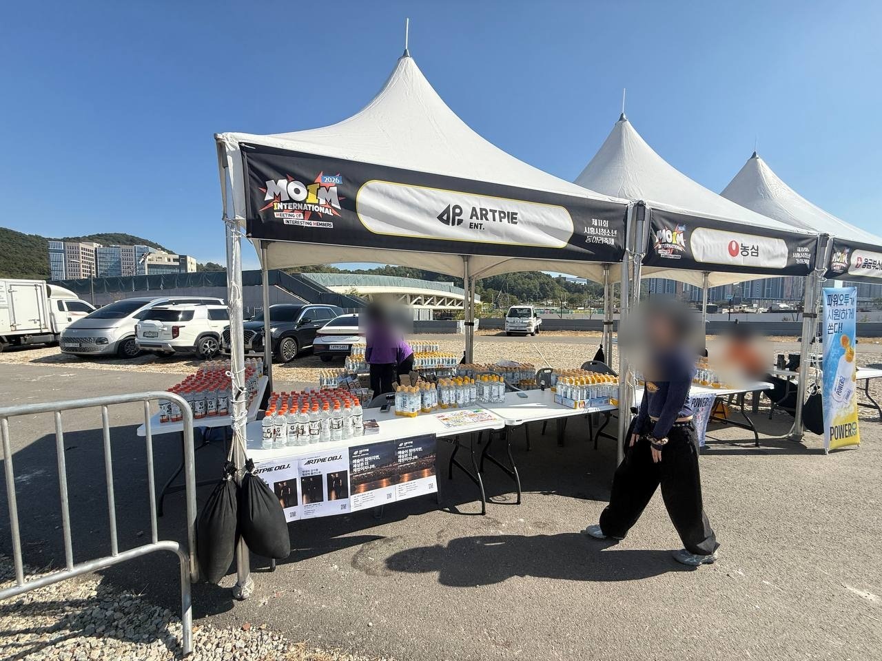 MOIM INTERNATIONAL과 ARTPIE ENT. 현수막이 걸린 흰 천막 아래 테이블에 음료가 줄지어 놓여 있다. 인물 얼굴은 흐리게 처리했다