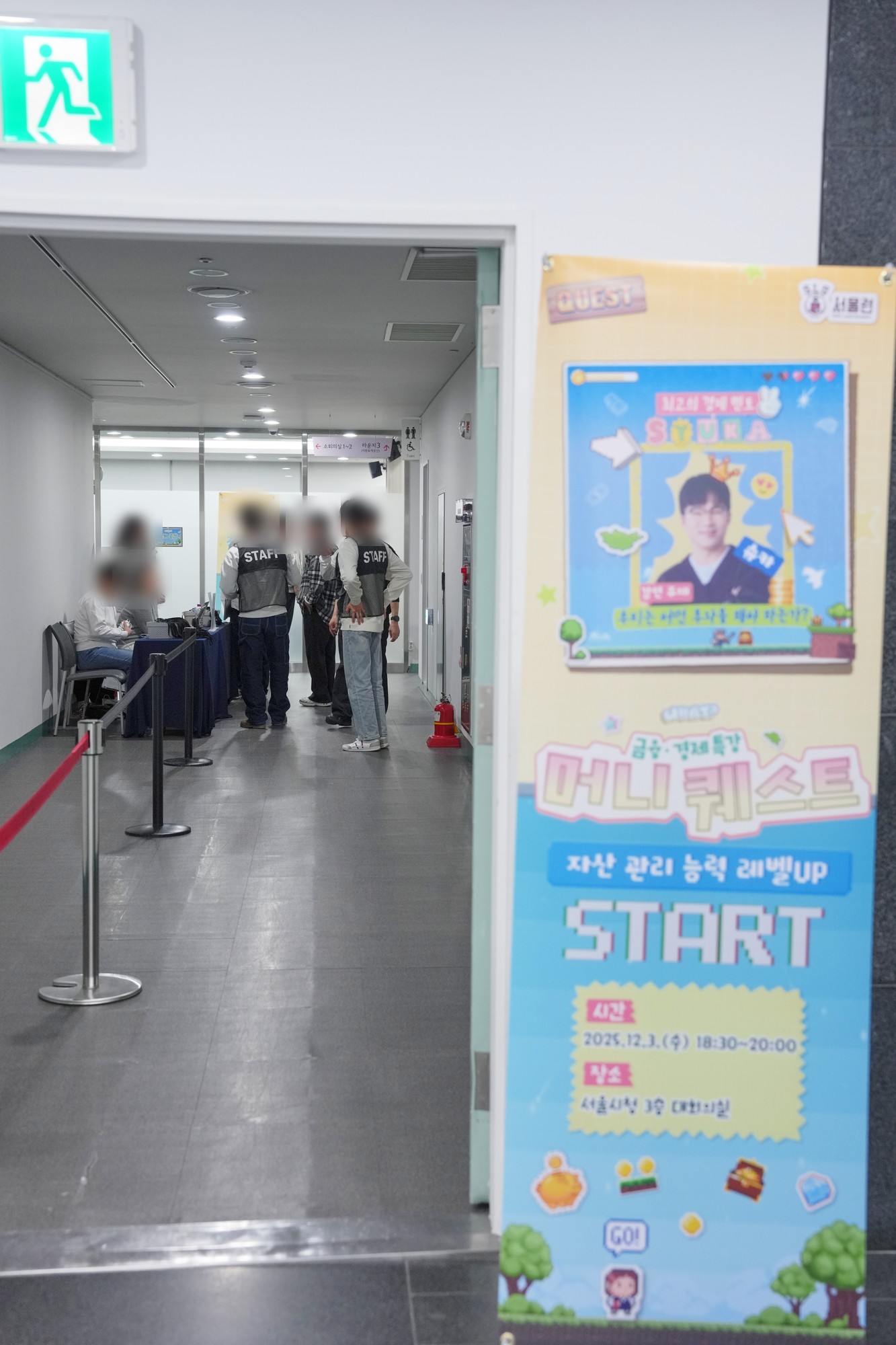 회의실 입구에 머니 퀘스트 배너가 서 있고 복도에 안전차단봉과 등록 테이블, STAFF 조끼를 입은 스태프들이 보인다. 인물 얼굴은 흐리게 처리했다