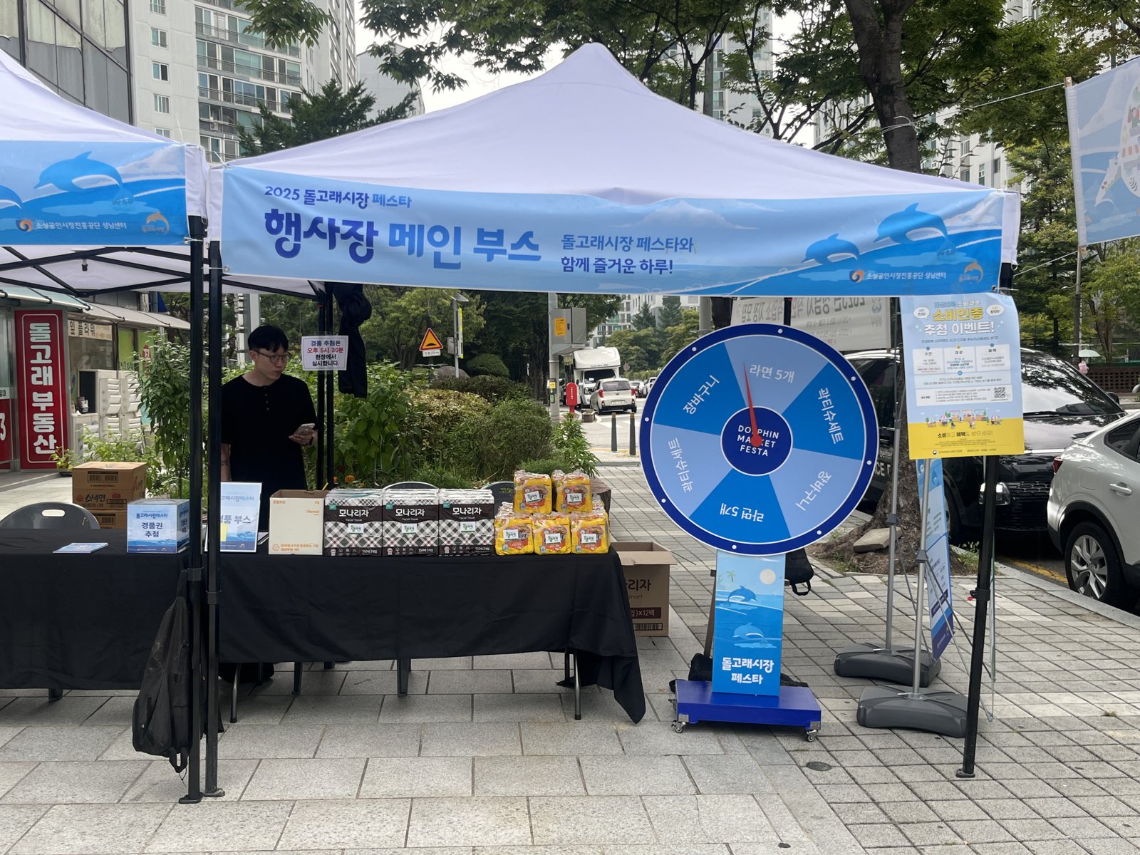 하늘색 캐노피 아래 대형 룰렛과 경품 상자가 놓인 테이블이 있고 스태프가 부스를 지키고 있다