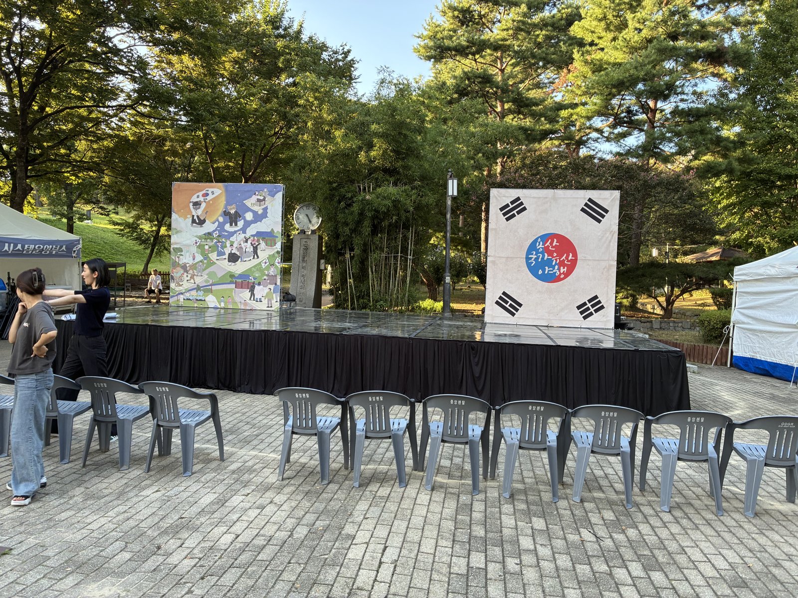 나무 그늘 아래 검은 단 무대 뒤로 태극 문양 패널과 그래픽 패널이 서 있고 앞에 의자가 놓여 있다