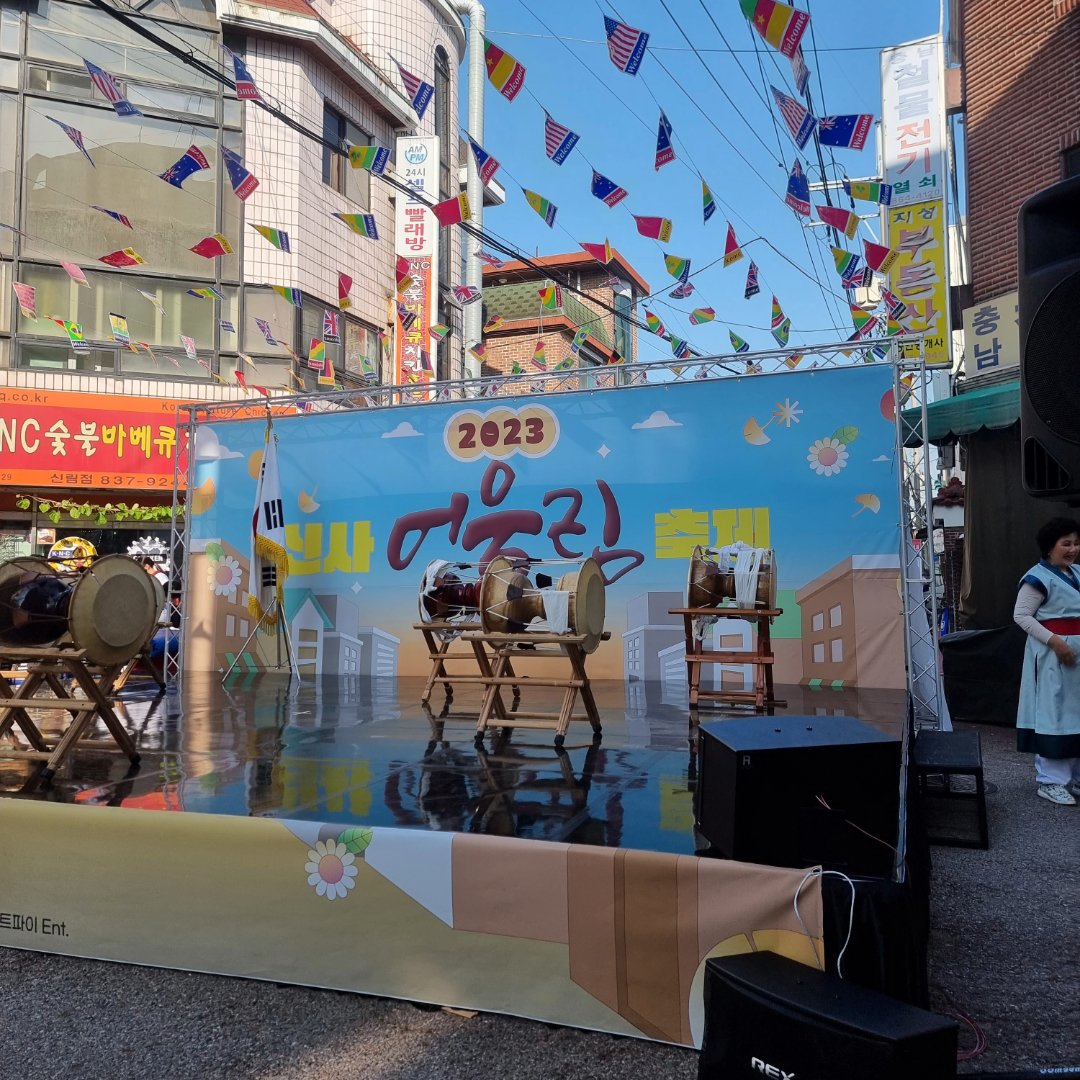 만국기가 걸린 시장 골목의 거리 무대를 멀리서 본 장면. 무대 위에 밴드가 있고 앞에 의자가 놓여 있다
