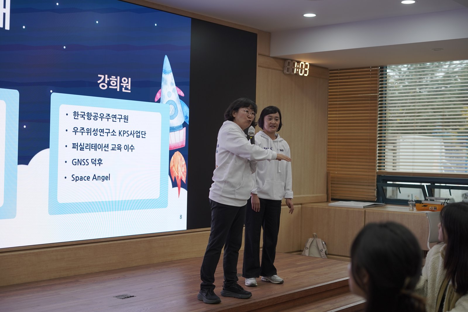 우주 그래픽 화면 앞 단상에서 흰 후드티를 입은 두 사람이 마이크를 들고 화면을 가리키며 소개하고 있다