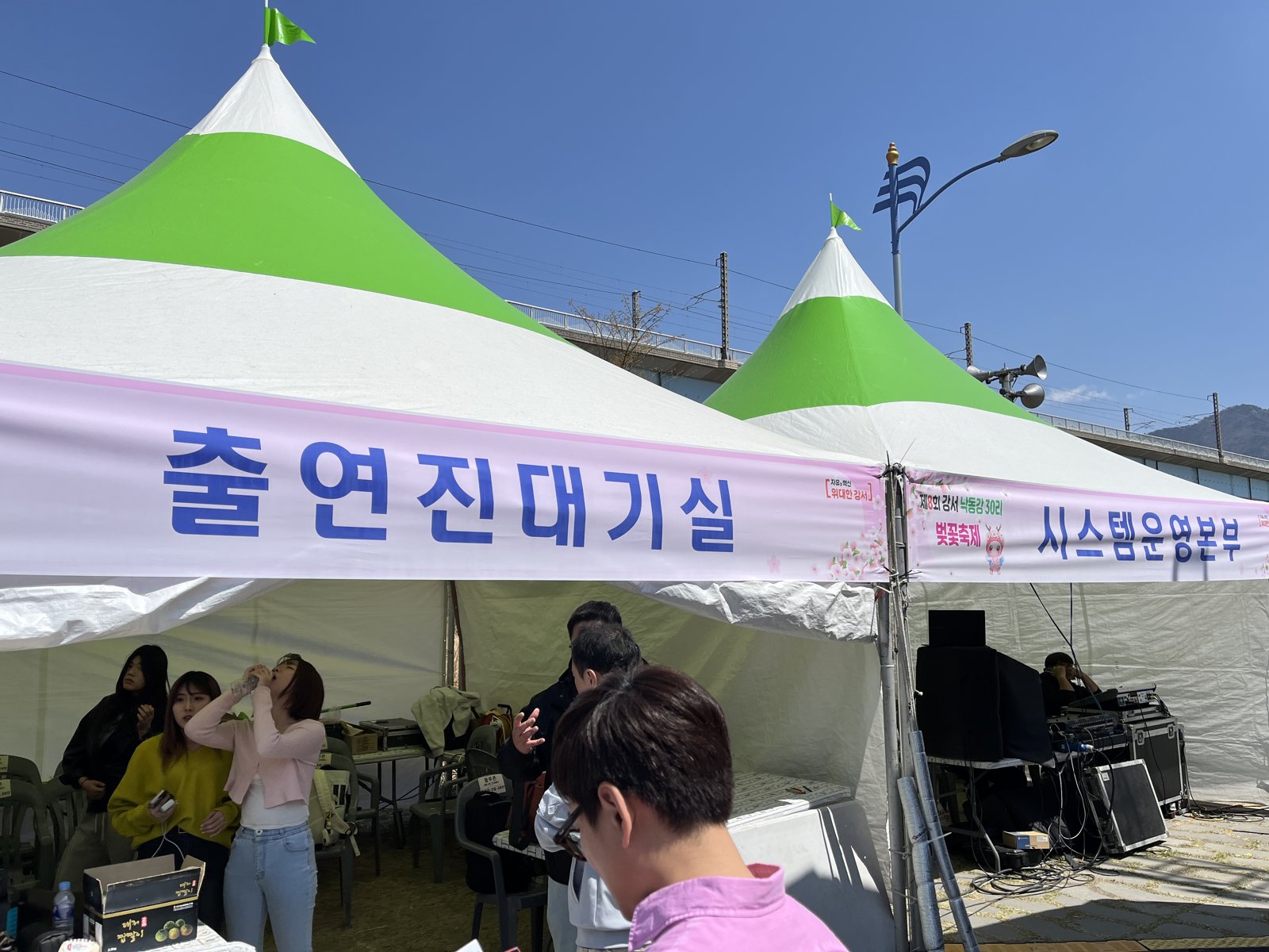 출연진대기실이라고 적힌 초록 흰색 천막 아래에서 출연자들이 공연을 준비하고 있고 옆 천막에는 시스템운영본부 현수막과 음향 장비가 보인다
